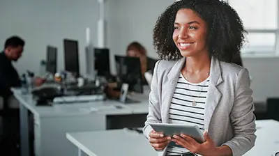 Driving Innovation Through the Digital Ecosystem of Insurance A woman in a horizontally striped shirt and a gray blazer looking ahead, while smiling and holding a tablet in her hands